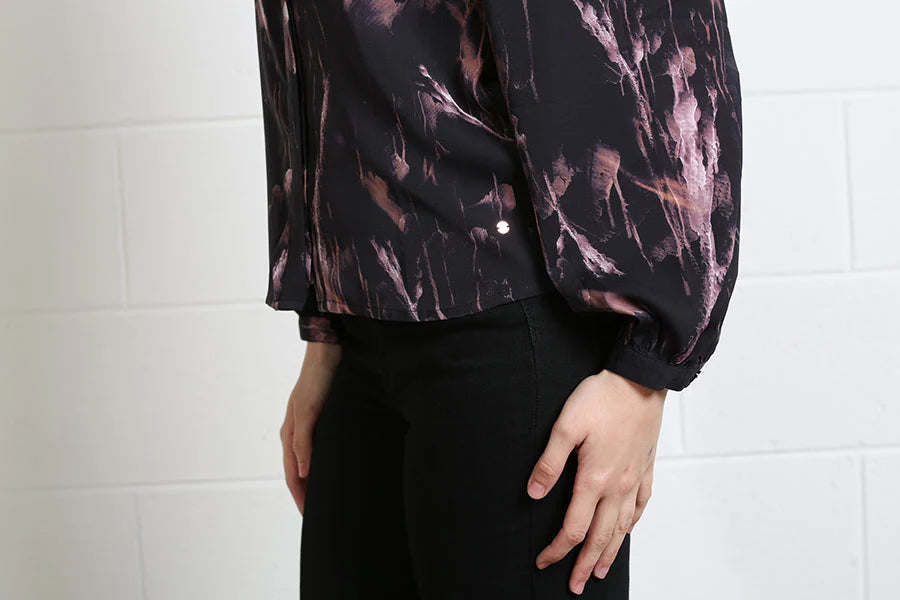 A person wearing a black long-sleeve blouse with a pink and brown abstract brushstroke pattern, paired with black pants, standing against a white brick wall.