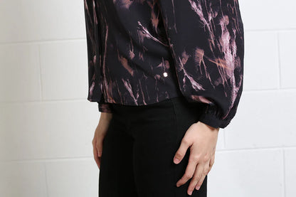 A person wearing a black long-sleeve blouse with a pink and brown abstract brushstroke pattern, paired with black pants, standing against a white brick wall.
