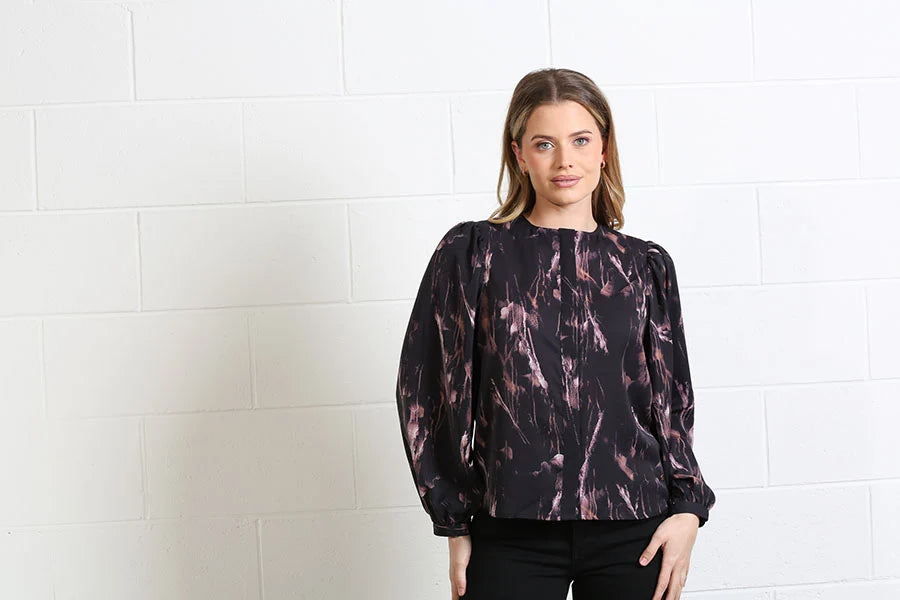 A woman modeling a long-sleeve blouse with a black and dusty rose abstract pattern, standing against a white brick wall.