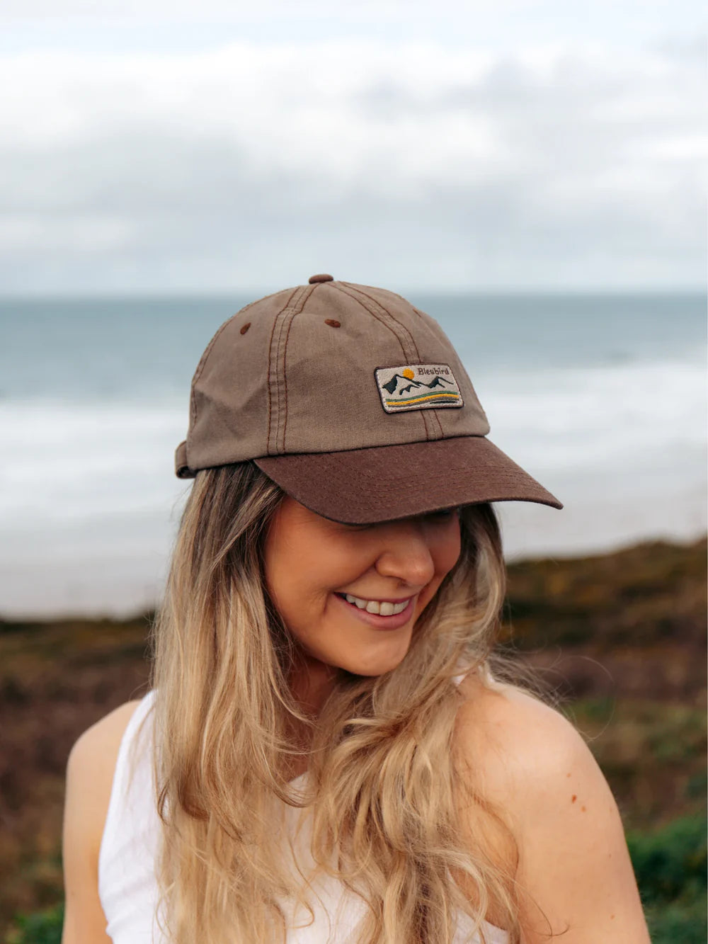 Woman wearing a brown cap with a logo, standing outdoors with a scenic background.