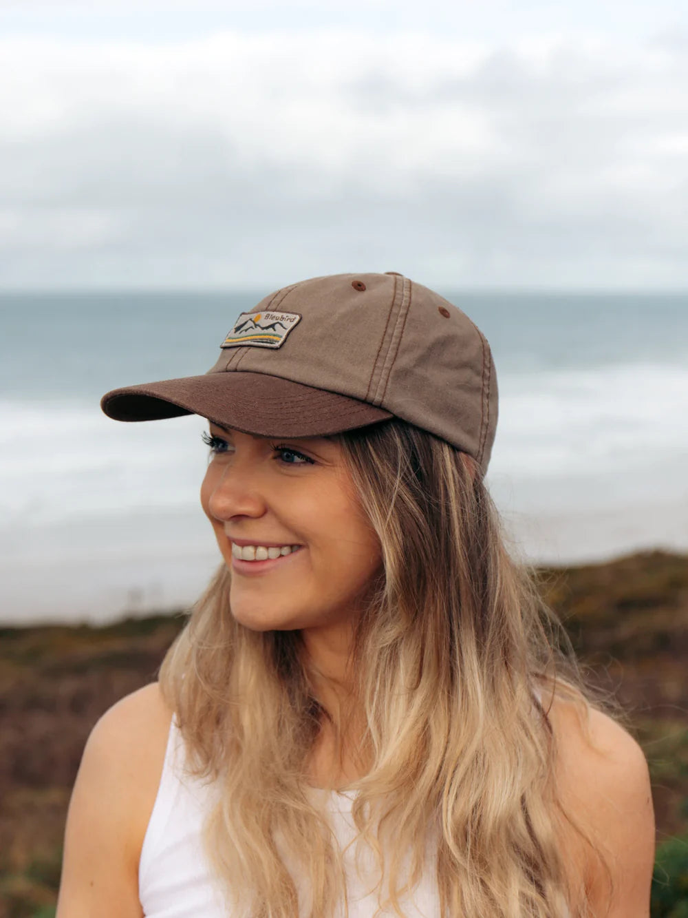 Woman wearing a brown cap with a patch, standing outdoors with a scenic background.
