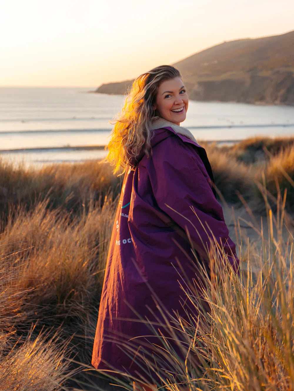 Woman in a purple coat standing in tall grass with a scenic background