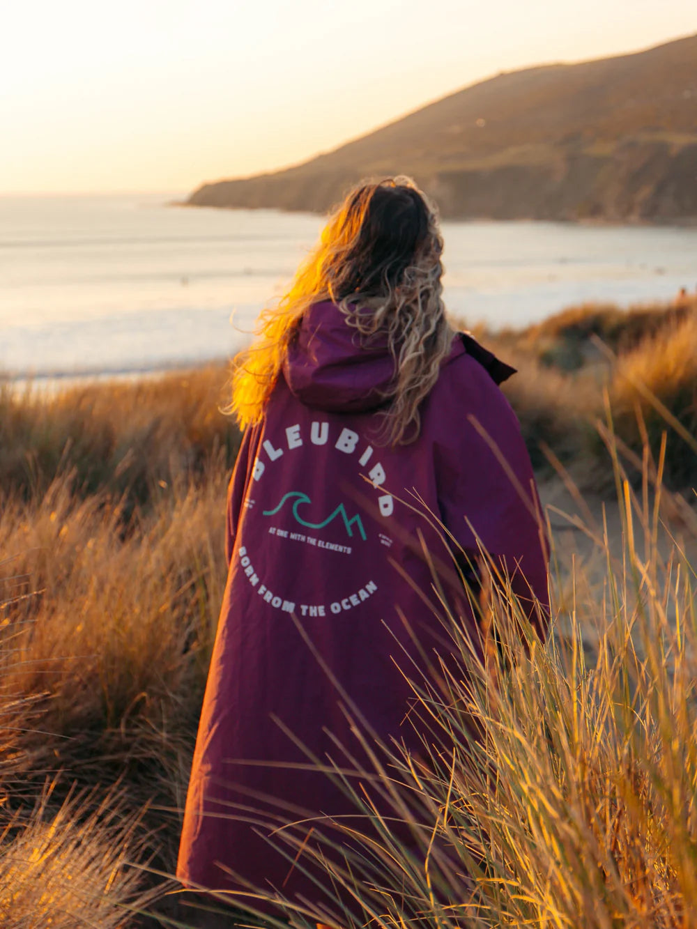 Person wearing a purple hoodie with branding, standing in tall grass by a scenic lakeside.