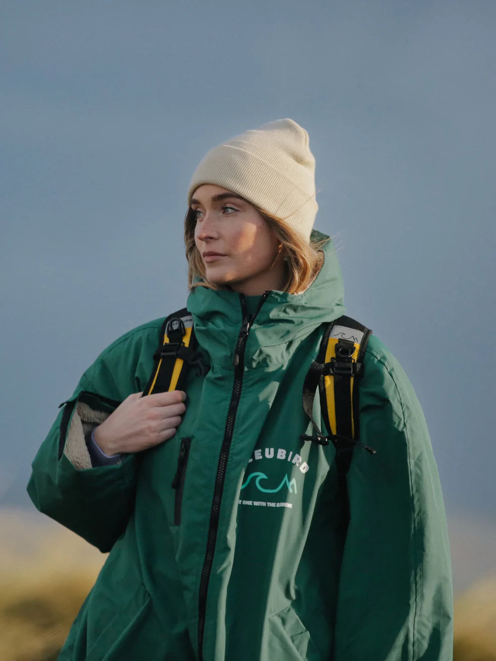 Person wearing a green jacket with a brand logo, standing outdoors against a blue sky.