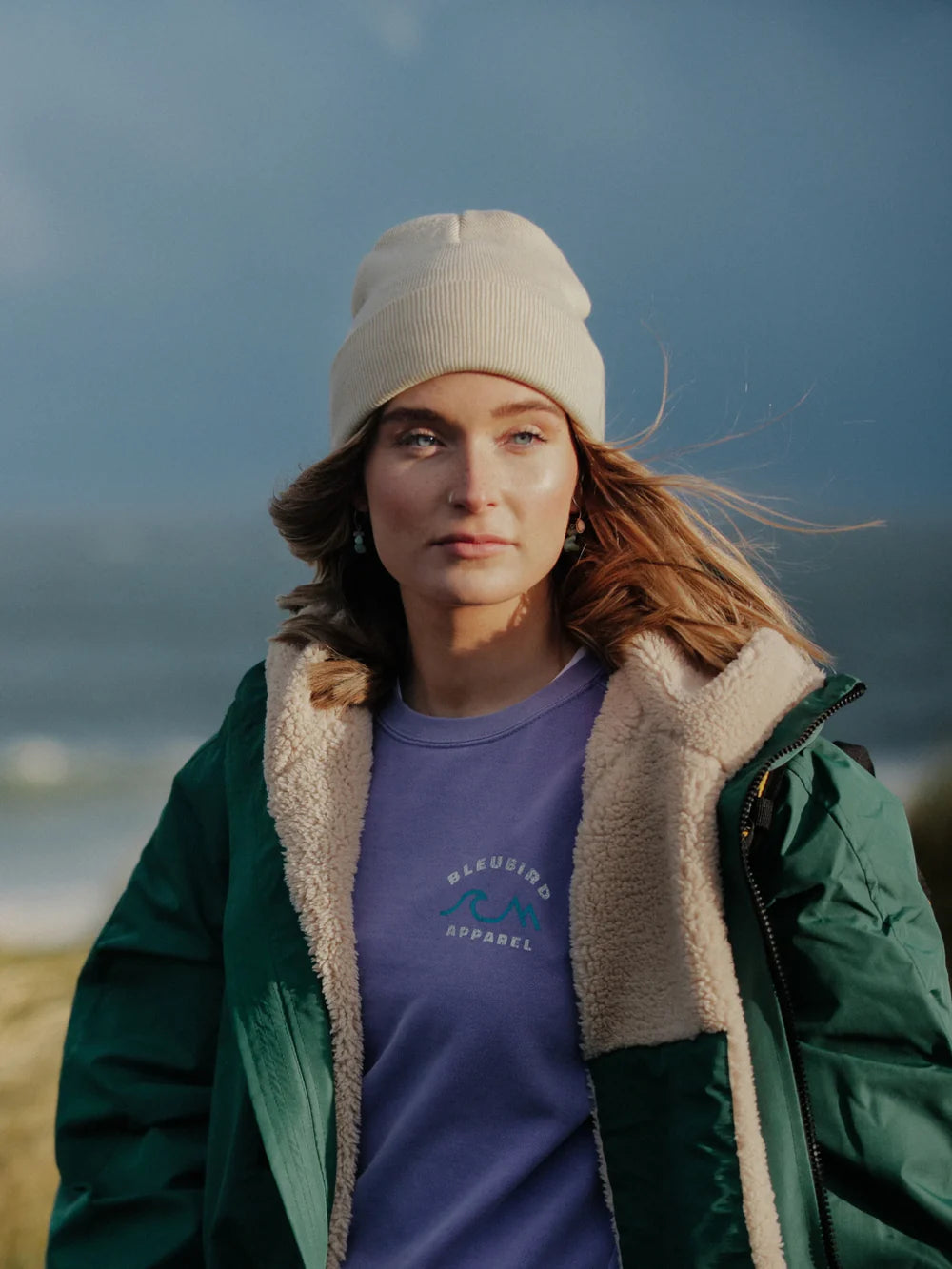 Woman wearing a green jacket and purple shirt with a logo, standing outdoors with a blue sky background.