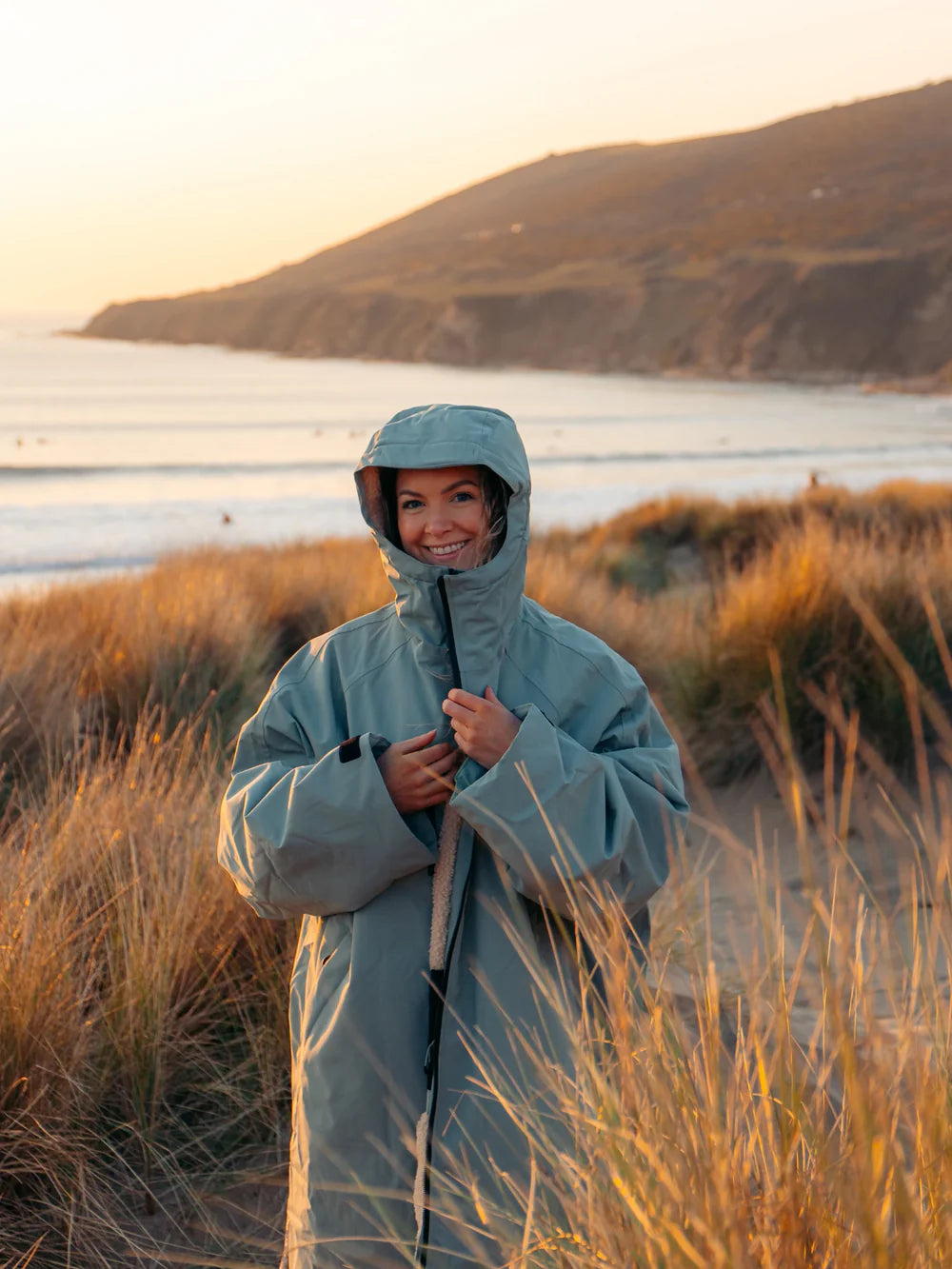 Person wearing a light blue hooded coat standing in tall grass by a body of water with mountains in the background.