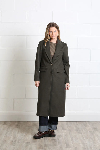 A woman stands in a studio wearing a long dark olive green coat over a taupe top, cuffed blue jeans, and brown shoes, against a white brick wall and light wood floor.