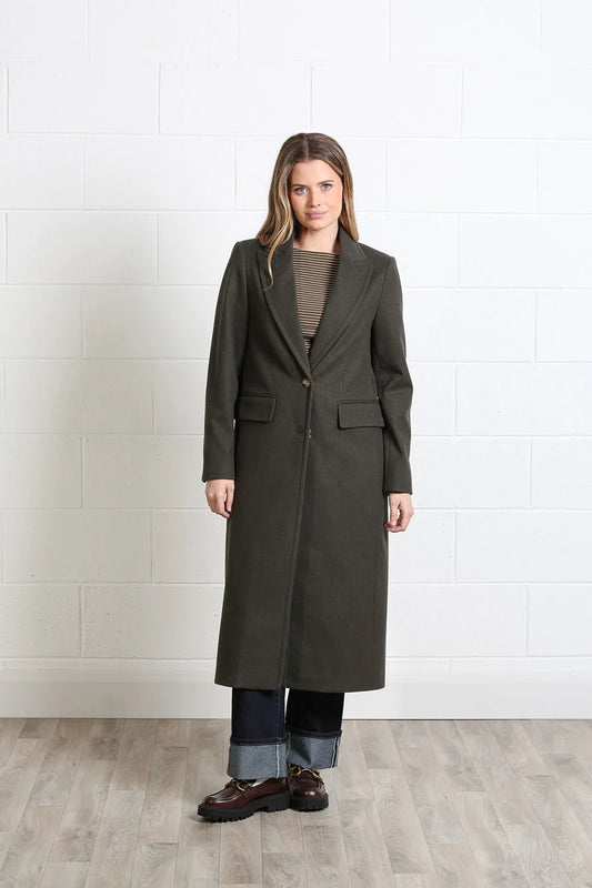 A woman stands in a studio wearing a long dark olive green coat over a taupe top, cuffed blue jeans, and brown shoes, against a white brick wall and light wood floor.