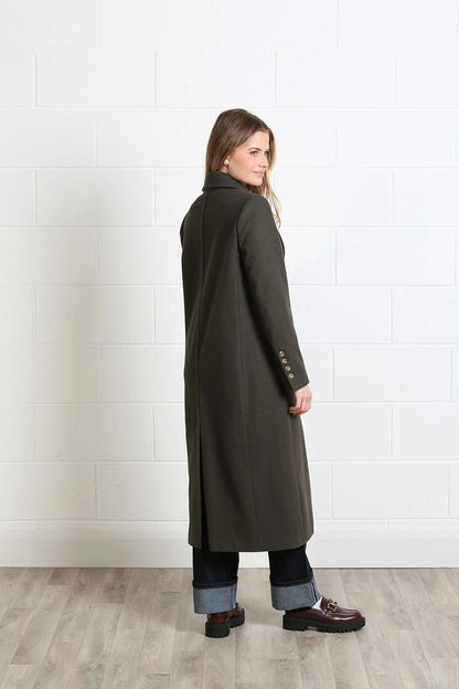A woman wearing a long dark olive green coat, cuffed blue jeans, and brown loafers, standing in profile against a white brick wall and light wood floor.