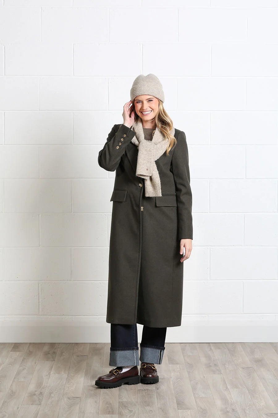 A woman stands in a studio wearing a long dark olive-brown coat, a light beige knit beanie, a matching knit scarf, dark jeans, and brown loafers against a white brick wall and light wood floor.