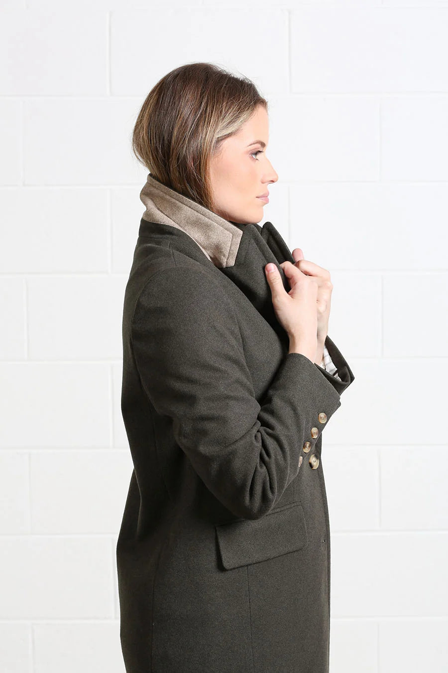 A woman shown in side profile wearing a dark olive green wool coat with a contrasting beige suede collar, standing against a white brick wall.