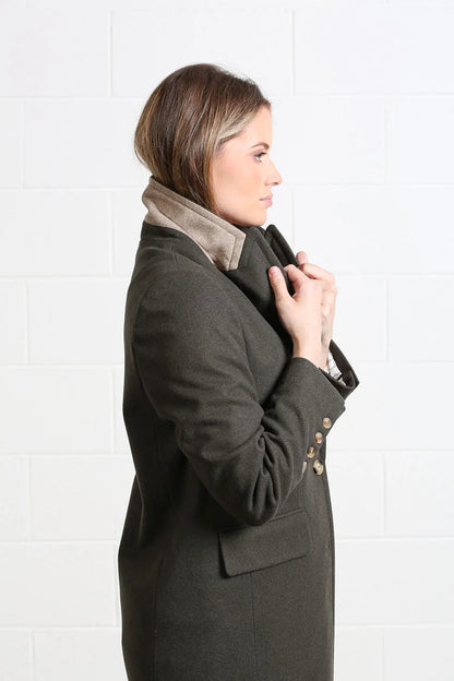 A woman shown in side profile wearing a dark olive green wool coat with a contrasting beige suede collar, standing against a white brick wall.