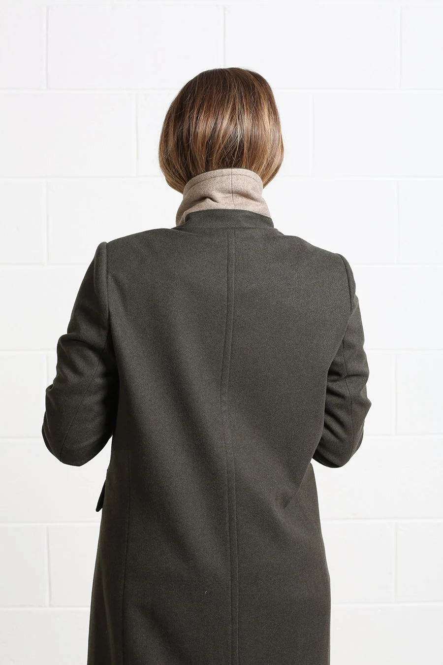 A person viewed from behind wearing a dark charcoal gray coat over a beige turtleneck, standing against a white tiled wall.