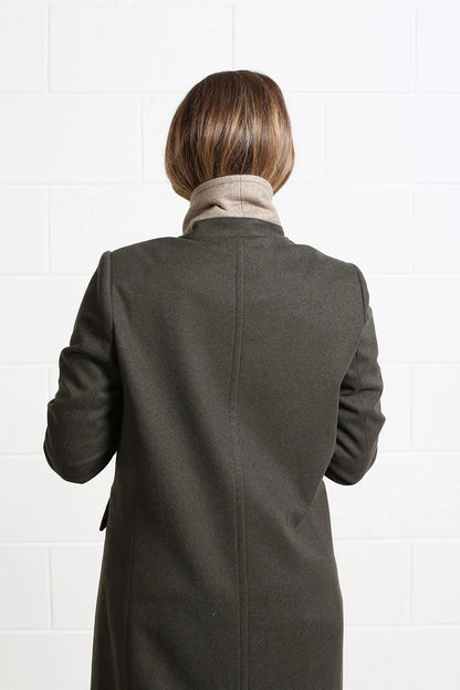 A person viewed from behind wearing a dark charcoal gray coat over a beige turtleneck, standing against a white tiled wall.