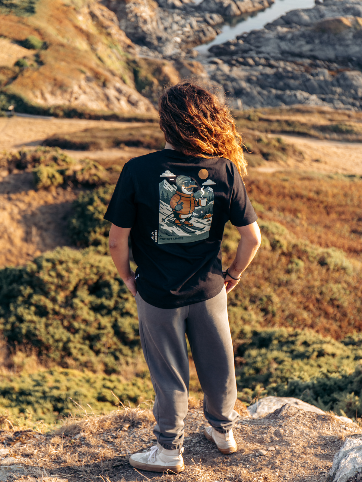 A person standing outdoors on a rocky hillside wearing a black graphic T-shirt with a large illustrated turtle design on the back, gray sweatpants, and white sneakers, looking out over a mountain landscape.