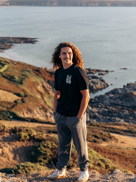 A man with long curly hair stands outdoors on a rocky coastal cliff wearing a black t-shirt with a small graphic on the chest, gray sweatpants, and white sneakers, with the sea and shoreline in the background.