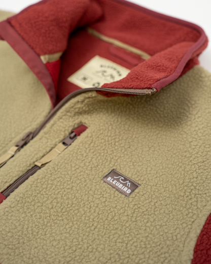 Close-up of a fleece pullover vest with a light olive-beige exterior, deep red interior lining, and a small black BLUBJORD logo patch on the chest.