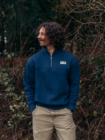 Man wearing a blue pullover with a logo standing in front of leafless branches.