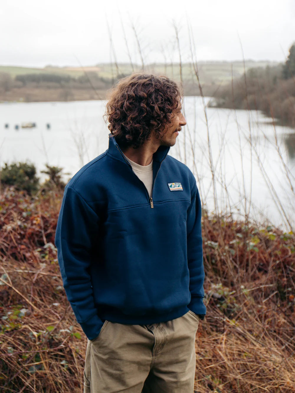 Man wearing a blue pullover with a logo standing by a lake