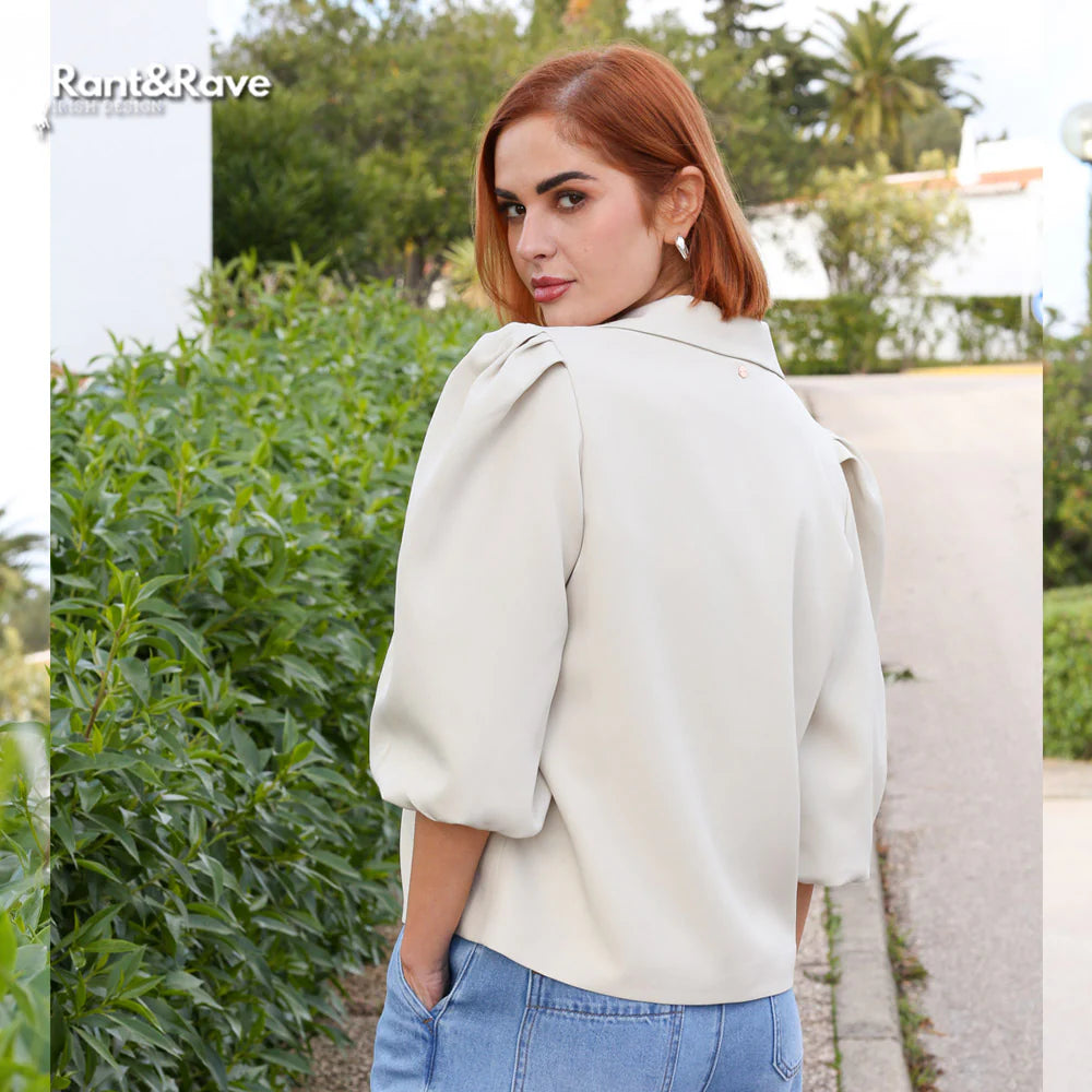 Woman wearing a white coat and blue jeans standing outdoors with greenery in the background