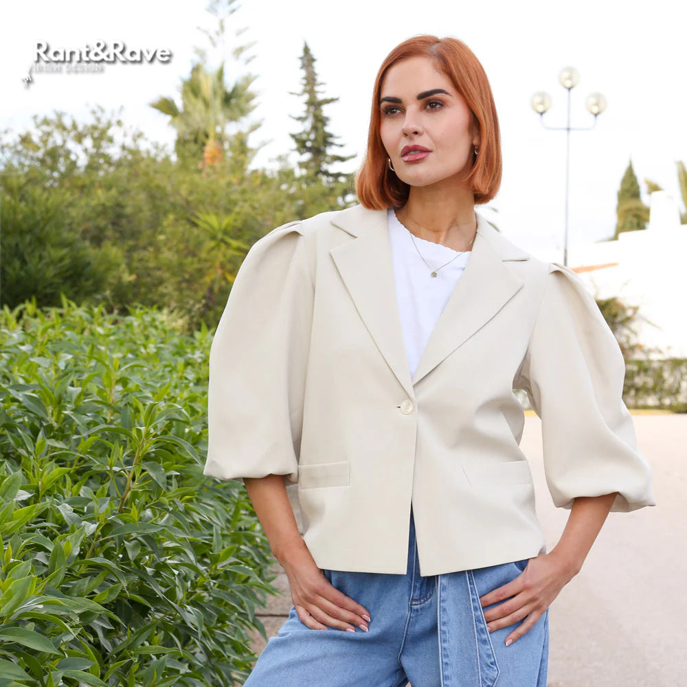 Woman wearing a beige blazer and blue jeans standing outdoors with greenery in the background