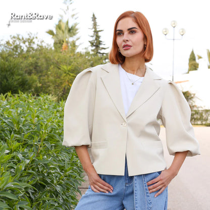 Woman wearing a beige blazer and blue jeans standing outdoors with greenery in the background