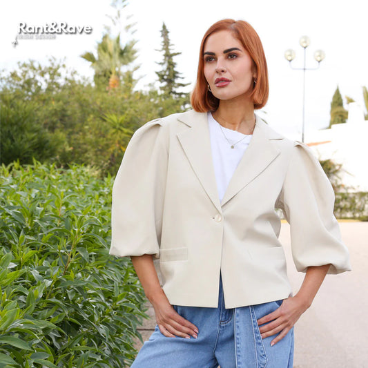 Woman wearing a beige blazer and blue jeans standing outdoors with greenery in the background