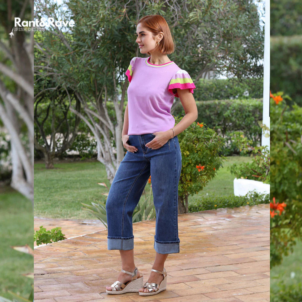 Woman wearing a colorful top and blue jeans standing on a wooden deck with greenery in the background