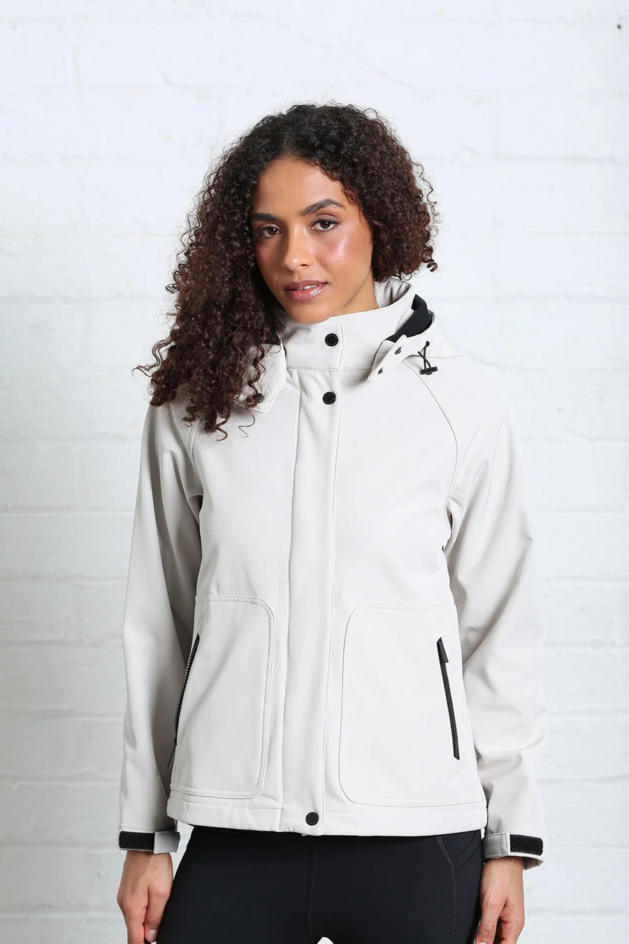 A woman with curly dark hair wearing a light gray hooded jacket with black trim, standing against a white brick wall.