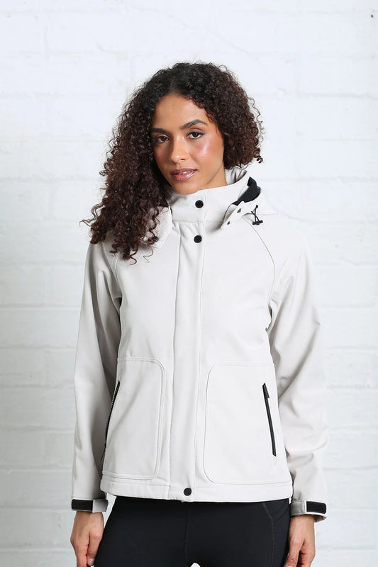 A woman with curly dark hair wearing a light gray hooded jacket with black trim, standing against a white brick wall.