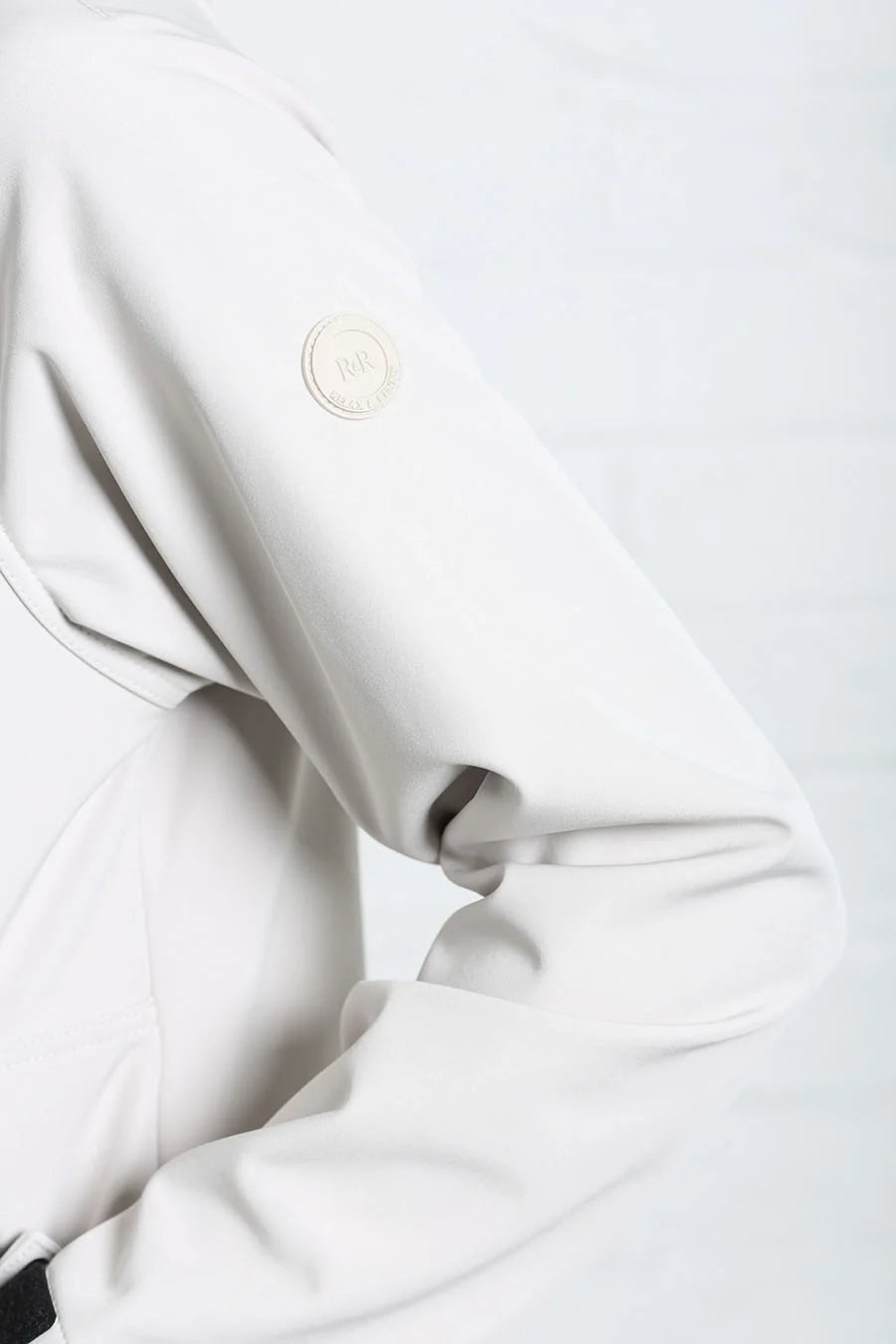 Close-up of a light gray or off-white jacket sleeve with a circular metallic emblem on the upper arm, set against a matching light gray background.