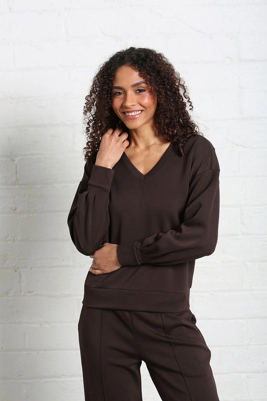 A woman with curly dark hair wearing a dark brown long-sleeve V-neck sweatshirt and matching dark brown sweatpants, standing against a white painted brick wall.