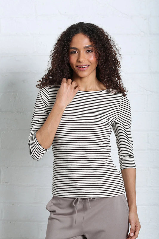 A woman with curly brown hair wearing a black and white horizontally striped three-quarter sleeve top and taupe drawstring sweatpants, standing against a white brick wall.