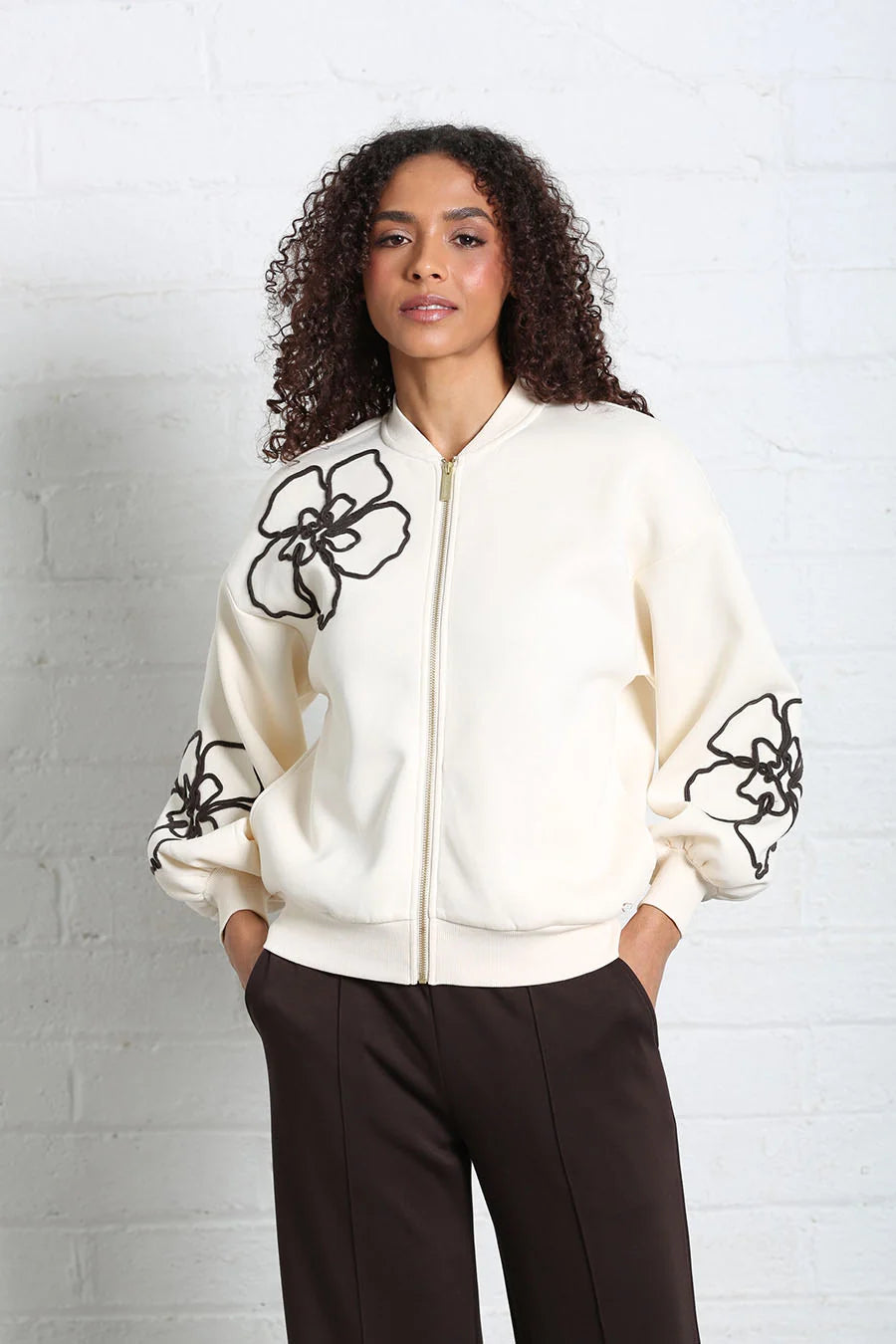 A woman with curly dark hair stands against a white brick wall wearing a cream zip-up bomber jacket with black floral embroidery on the chest and sleeves, paired with dark brown pants.
