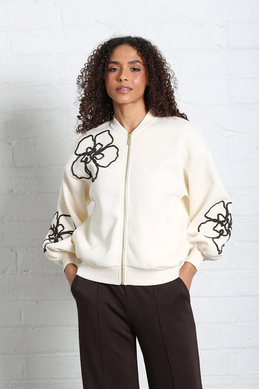 A woman with curly dark hair stands against a white brick wall wearing a cream zip-up bomber jacket with black floral embroidery on the chest and sleeves, paired with dark brown pants.
