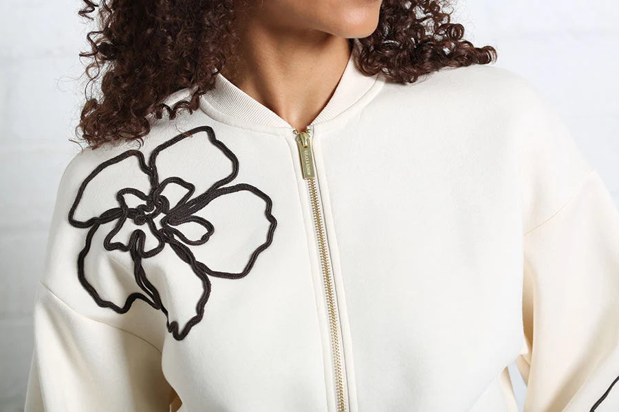 Close-up of a person wearing an off-white zip-up jacket with a large black floral outline embroidered on the chest and a gold zipper.