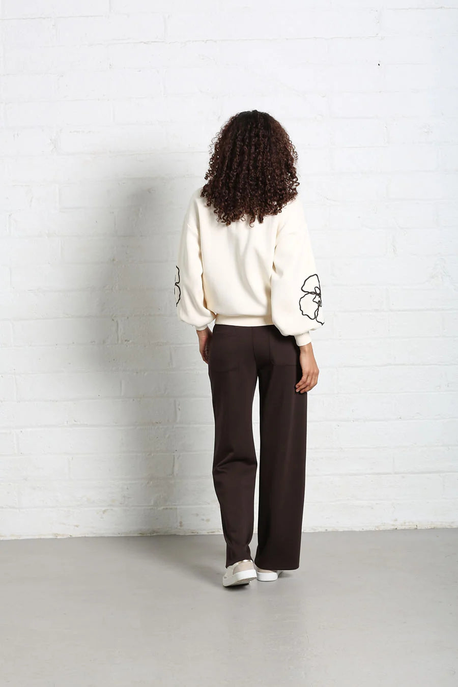 A model viewed from behind wearing a cream-colored sweatshirt with black graphic details on the sleeves and dark brown wide-leg trousers, standing against a white brick wall.