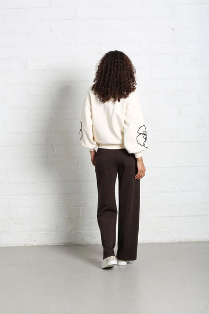 A model viewed from behind wearing a cream-colored sweatshirt with black graphic details on the sleeves and dark brown wide-leg trousers, standing against a white brick wall.