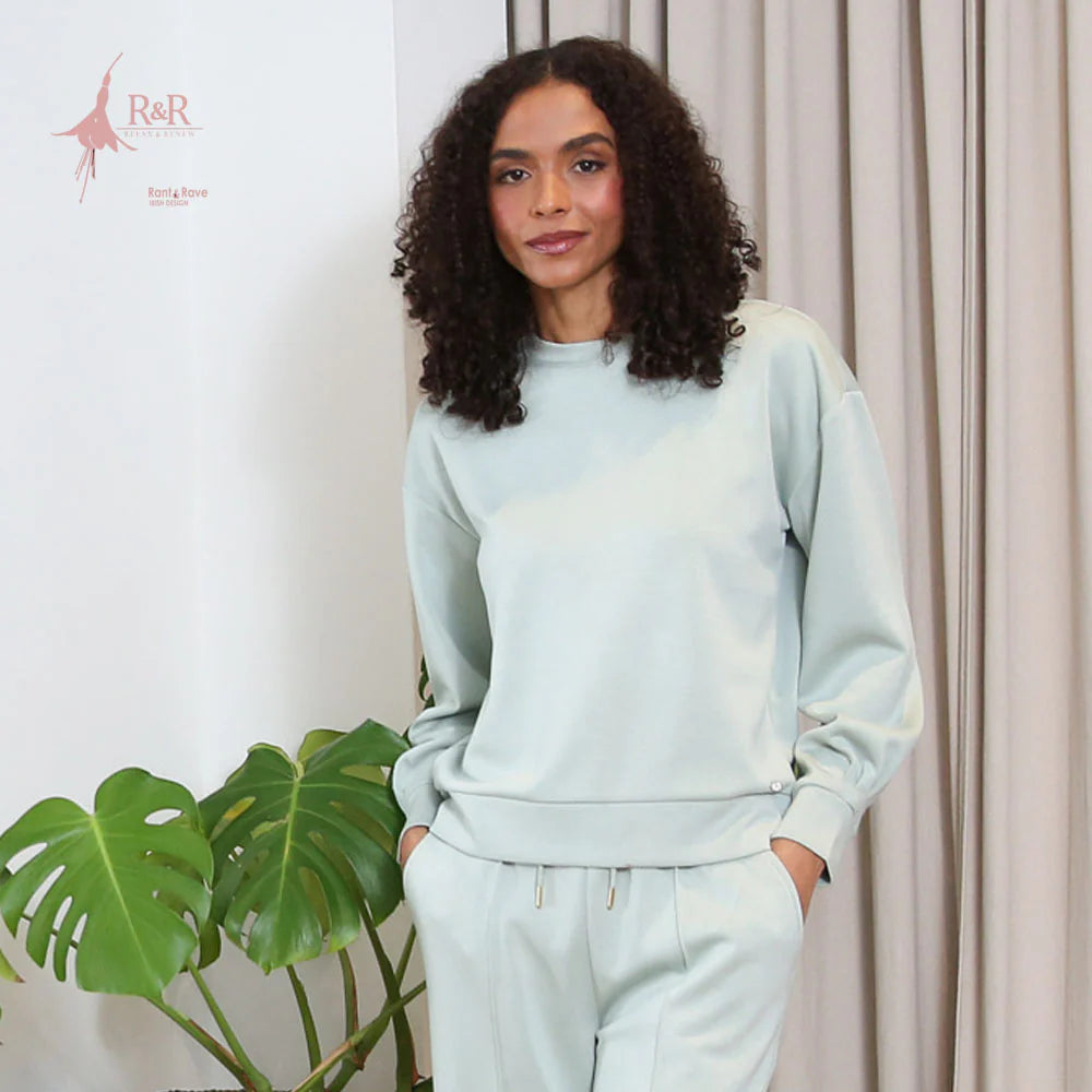 Woman wearing a light blue sweatshirt and pants standing next to a plant with a neutral background