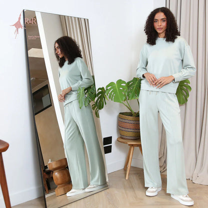 Woman in light green outfit standing next to a full-length mirror, with a plant in the background.