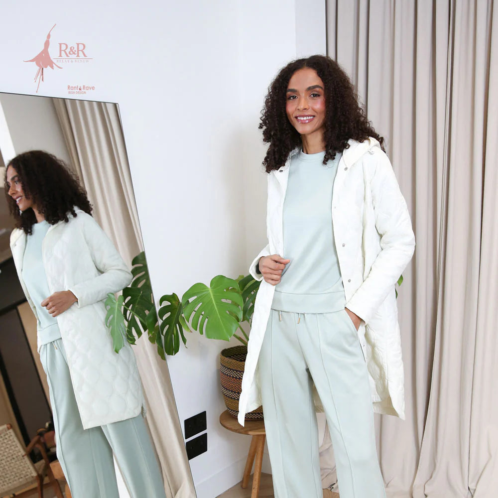 Woman in a light blue outfit with a white coat standing in a room with curtains and plants.