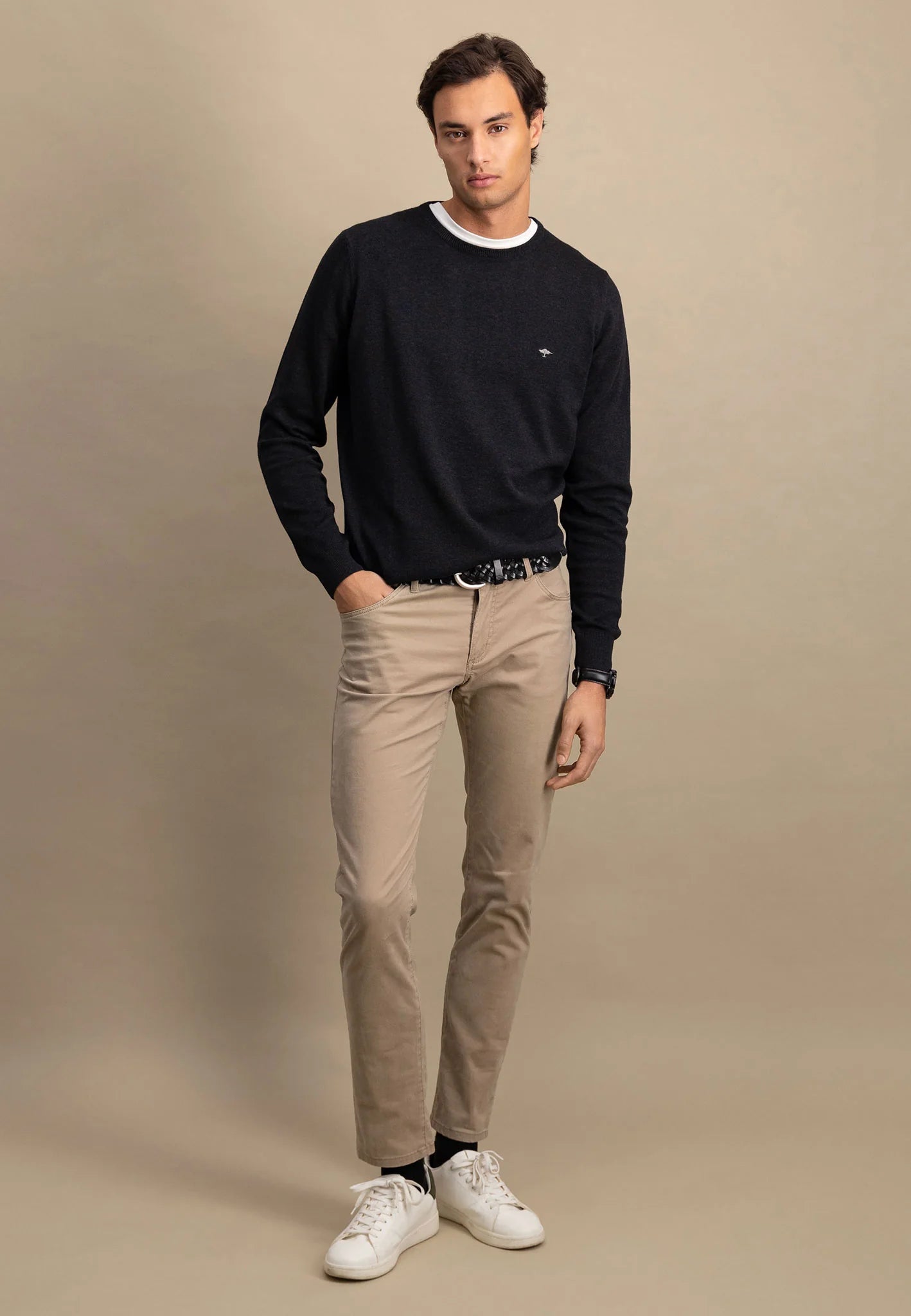 Man wearing a black sweater, beige pants, and white sneakers against a beige background