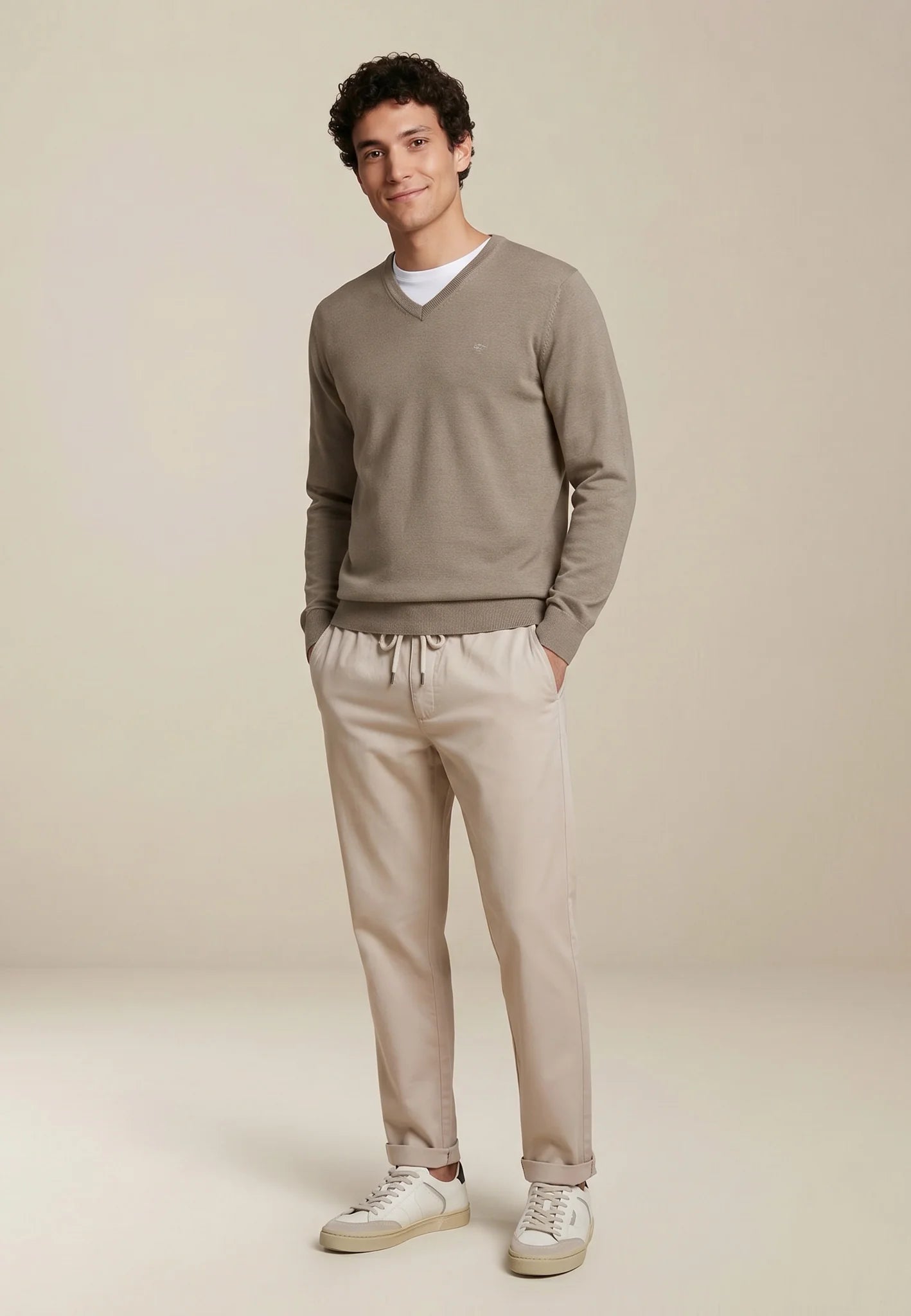 A man modeling a taupe v-neck sweater and beige drawstring trousers against a warm beige studio background.