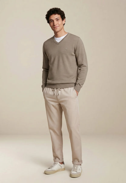 A man modeling a taupe v-neck sweater and beige drawstring trousers against a warm beige studio background.