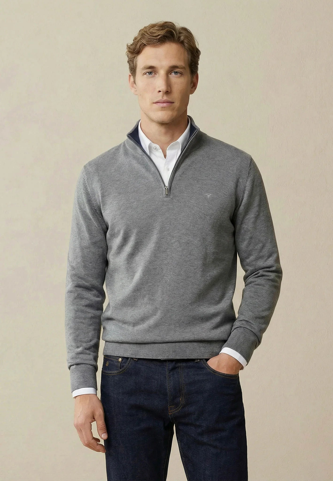 Man wearing a gray quarter-zip sweater over a white shirt with dark jeans on a beige background