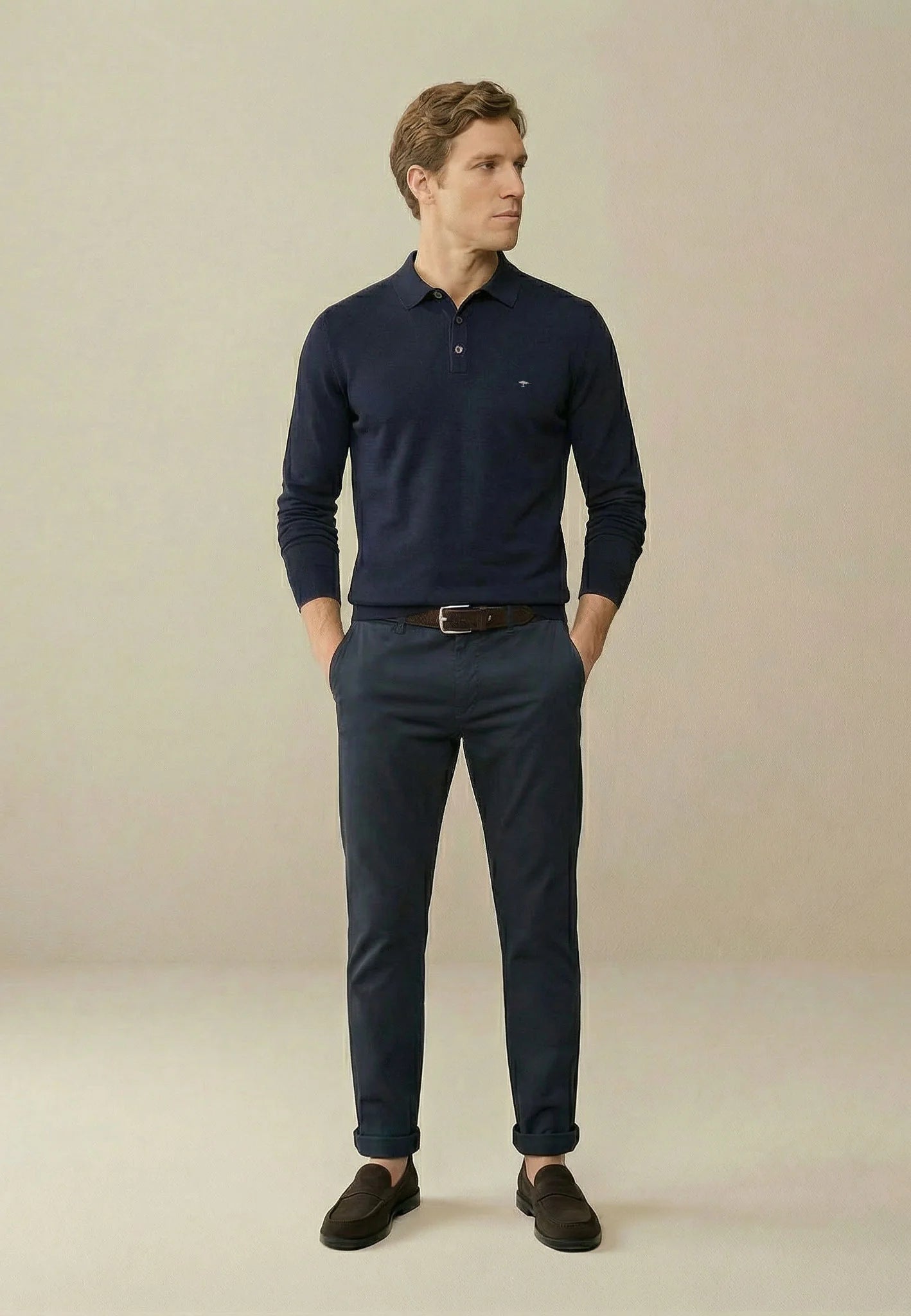 A man wearing a navy blue long-sleeve polo shirt, matching navy trousers, a brown belt, and dark brown slip-on shoes, standing against a plain beige studio background.