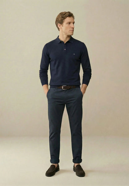 A man wearing a navy blue long-sleeve polo shirt, matching navy trousers, a brown belt, and dark brown slip-on shoes, standing against a plain beige studio background.