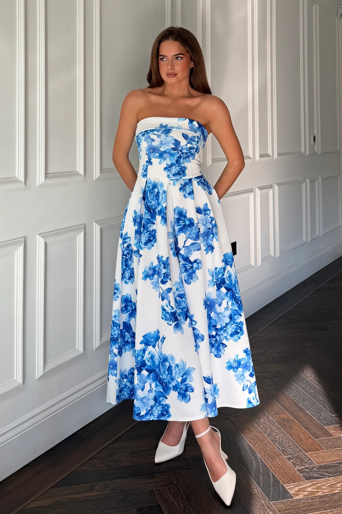 Woman wearing a strapless floral dress with blue flowers against a white paneled wall.