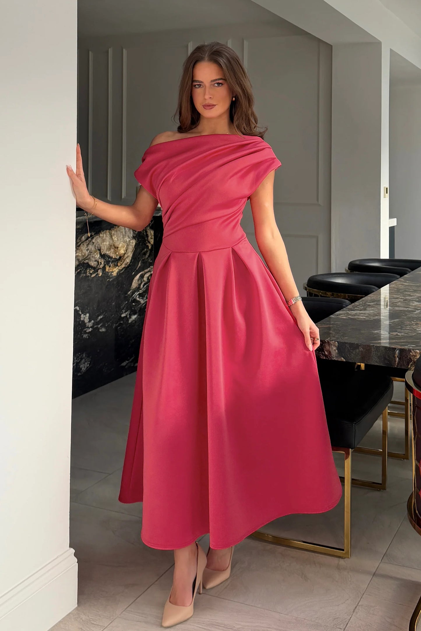 Woman in a pink off-shoulder dress standing in a modern interior setting.