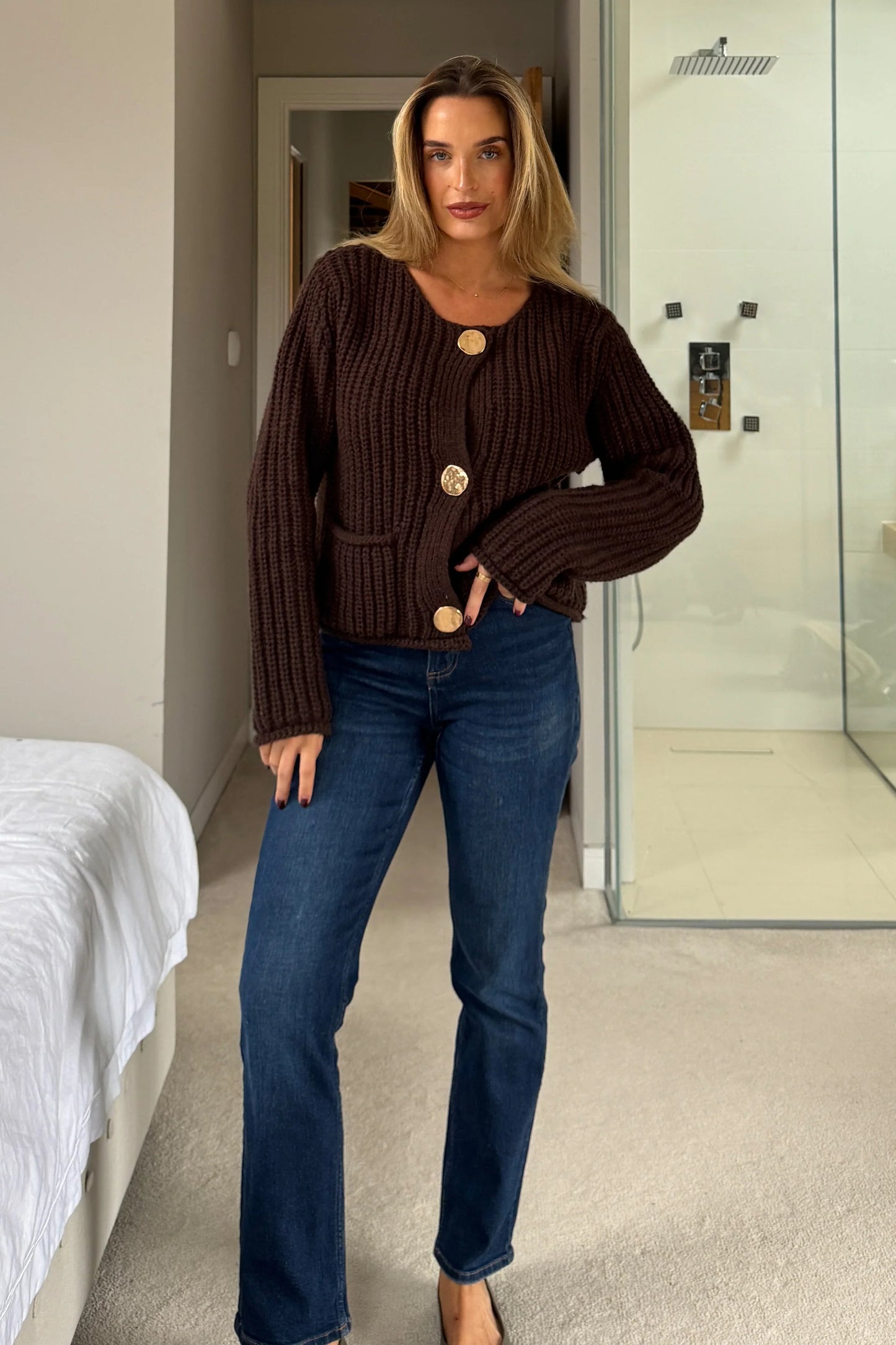 Woman wearing a brown ribbed sweater and blue jeans in a bedroom setting.